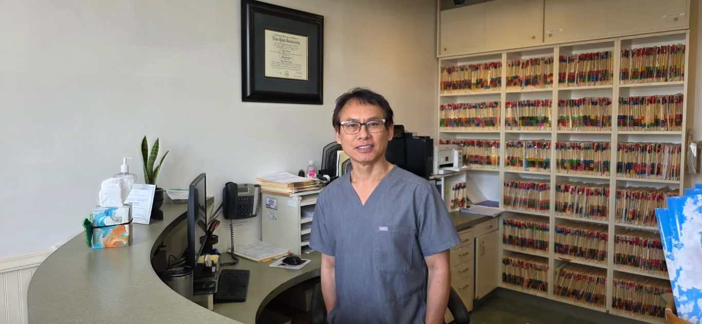 Dr. Kenneth Tran, DDS at the front desk of Peninsula Dentistry & Orthodontics in Huntington Beach, below his New York University dental diploma